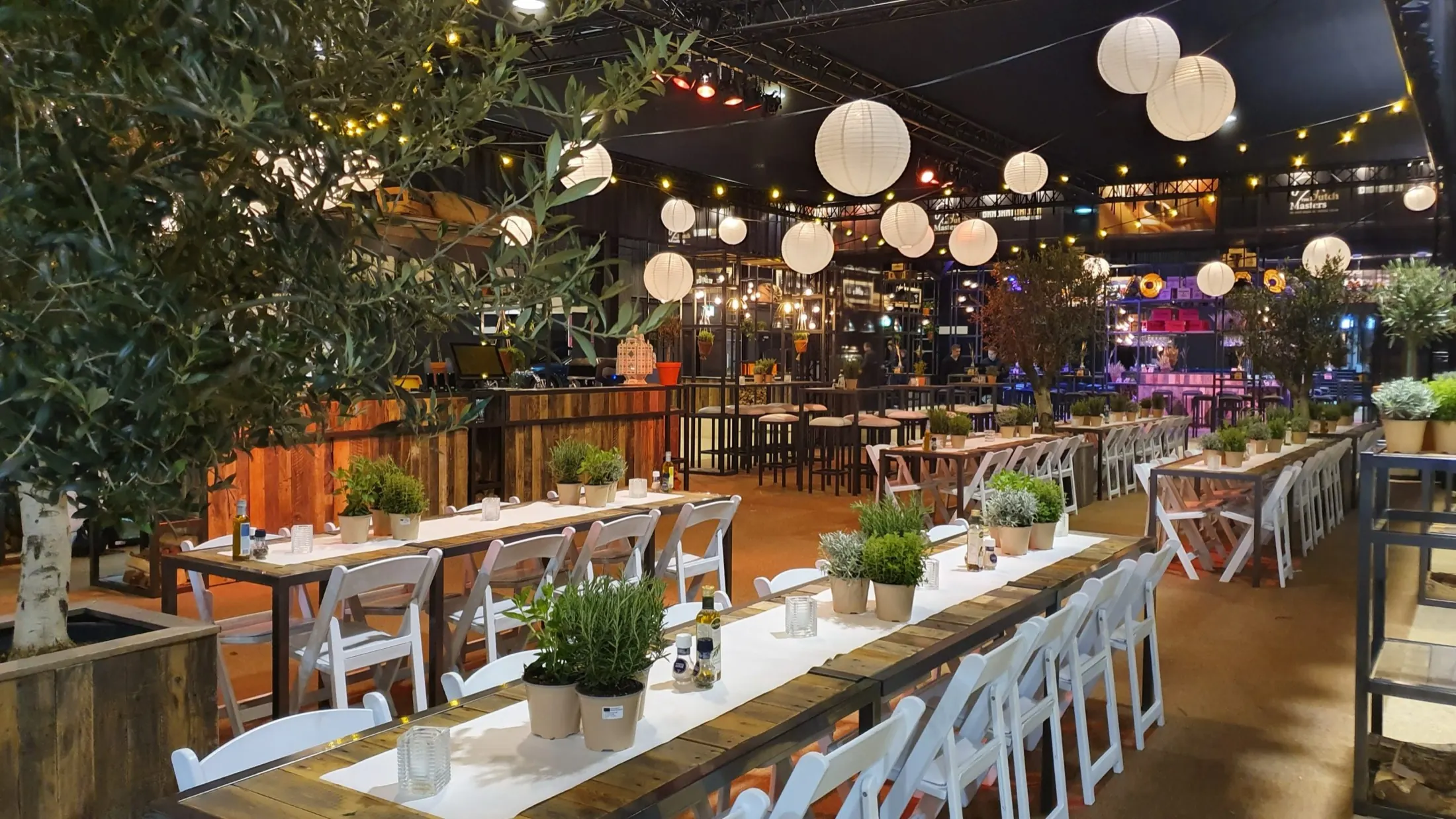 Restaurant met lange tafels, olijfbomen en papieren lampionnen