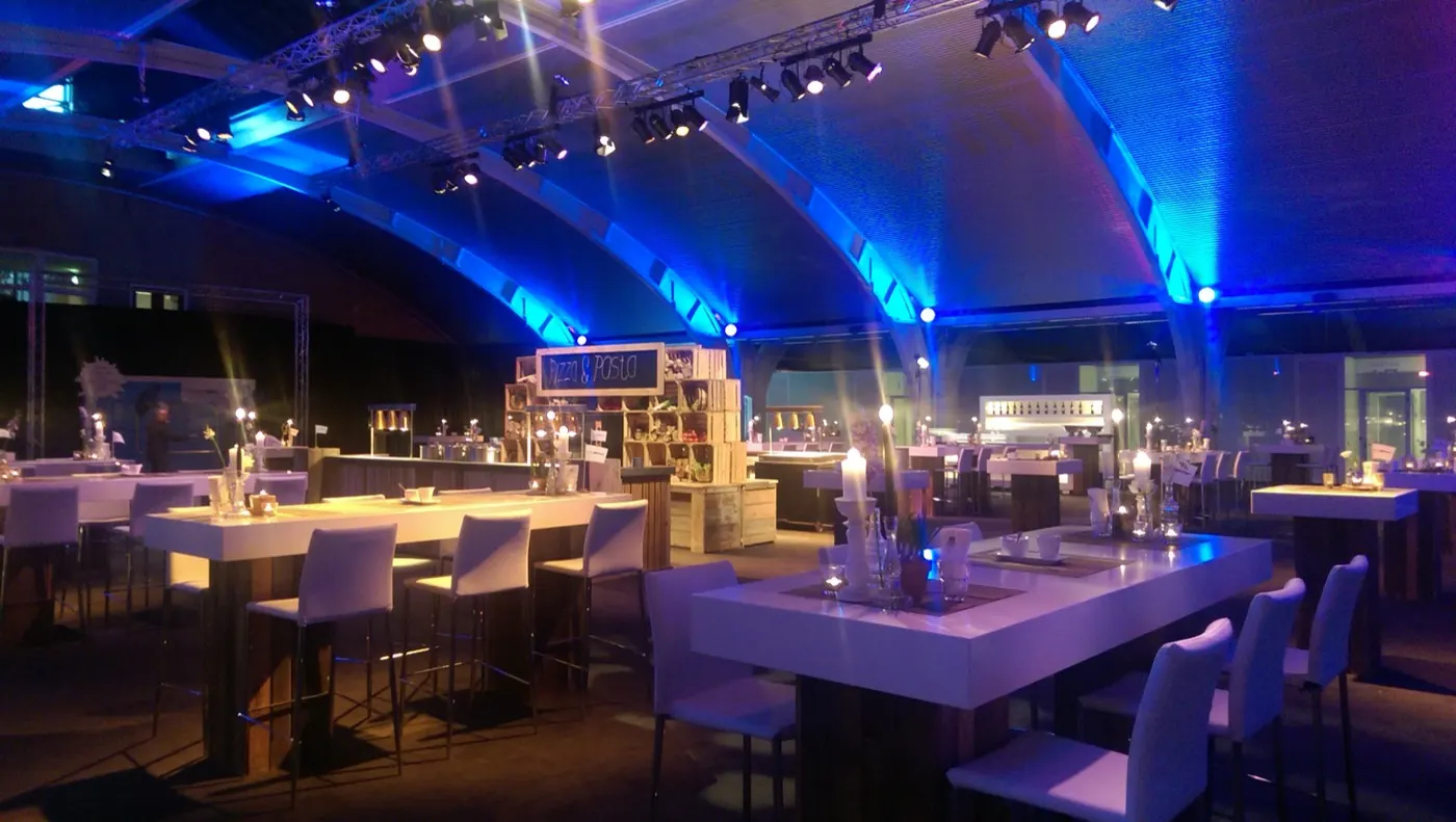 Zaal met witte statafels, kaarsen en blauw uplight tegen een gewelfd plafond